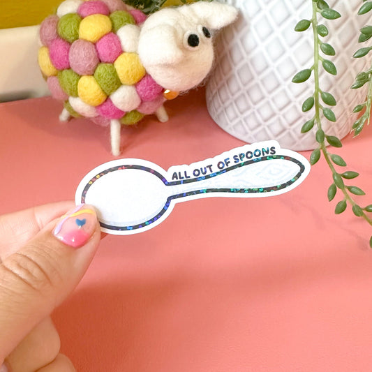 Hand holding a sticker with 'All Out of Spoons' text against a pink background with decorative items.