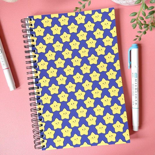 Notebook with yellow star pattern on a pink surface with pens and plants.