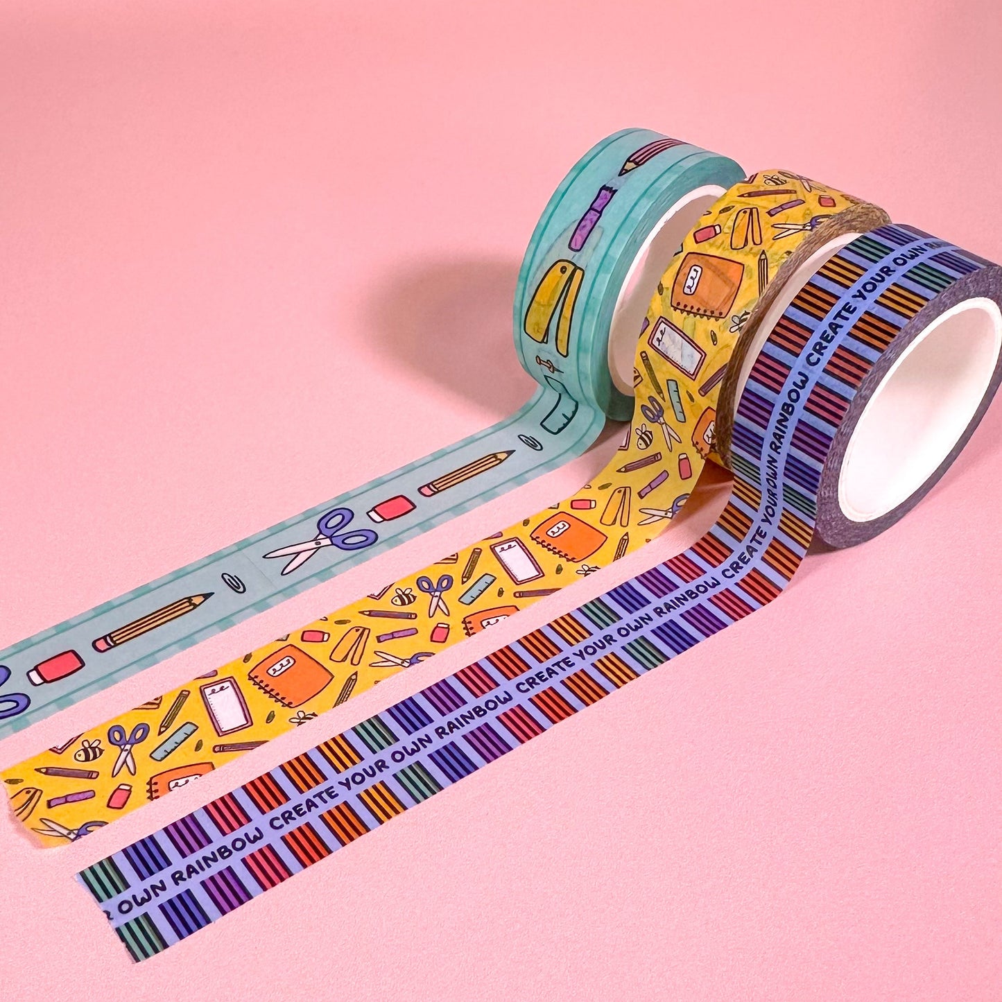 Three rolls of colorful washi tape with stationery-themed designs on a pink background.