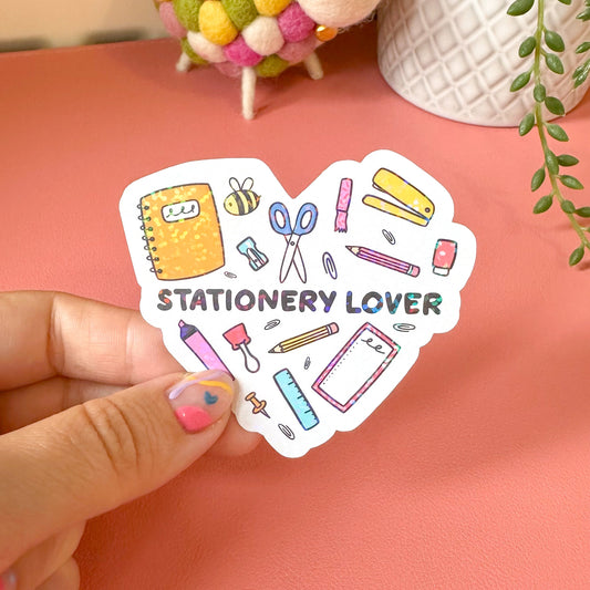 Heart-shaped sticker with stationery-themed illustrations held by a hand on a pink background.