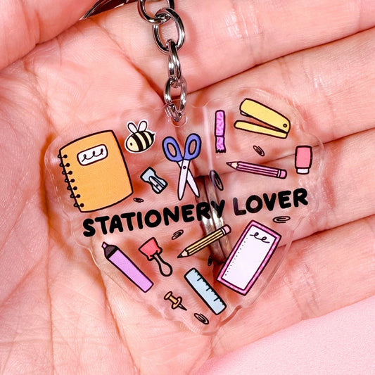 Clear keychain with stationery-themed charms and 'STATIONERY LOVER' text held in a hand against a pink background.