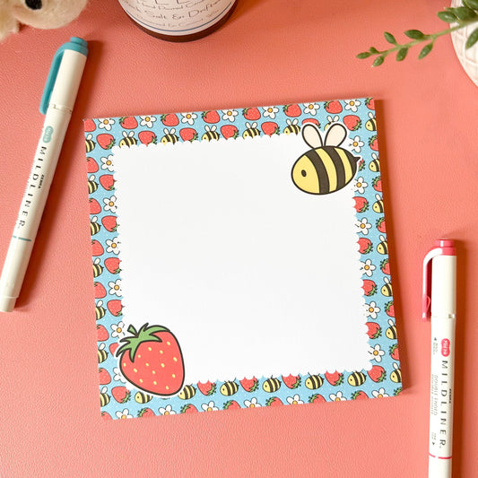 Notepad with strawberry and bee design on a pink surface with pens and a candle.