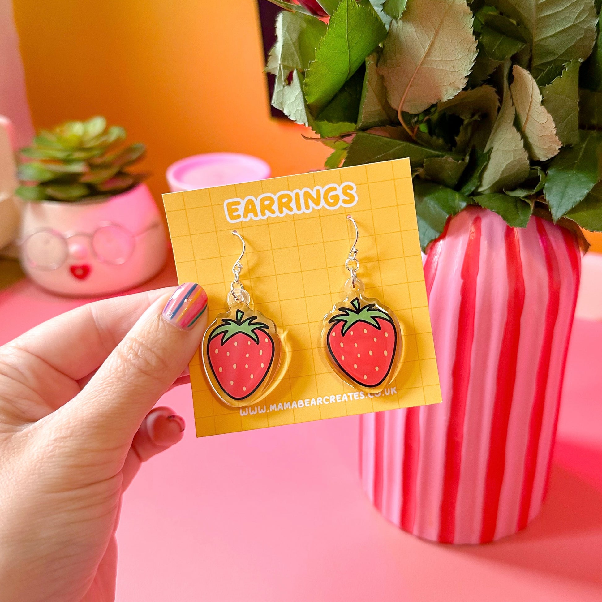 A pair of strawberry-shaped earrings on a yellow earring card, held in a person's hand with a decorative background.