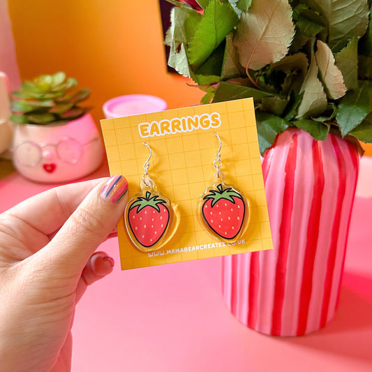 A pair of strawberry-shaped earrings on a yellow earring card, held in a person's hand with a decorative background.