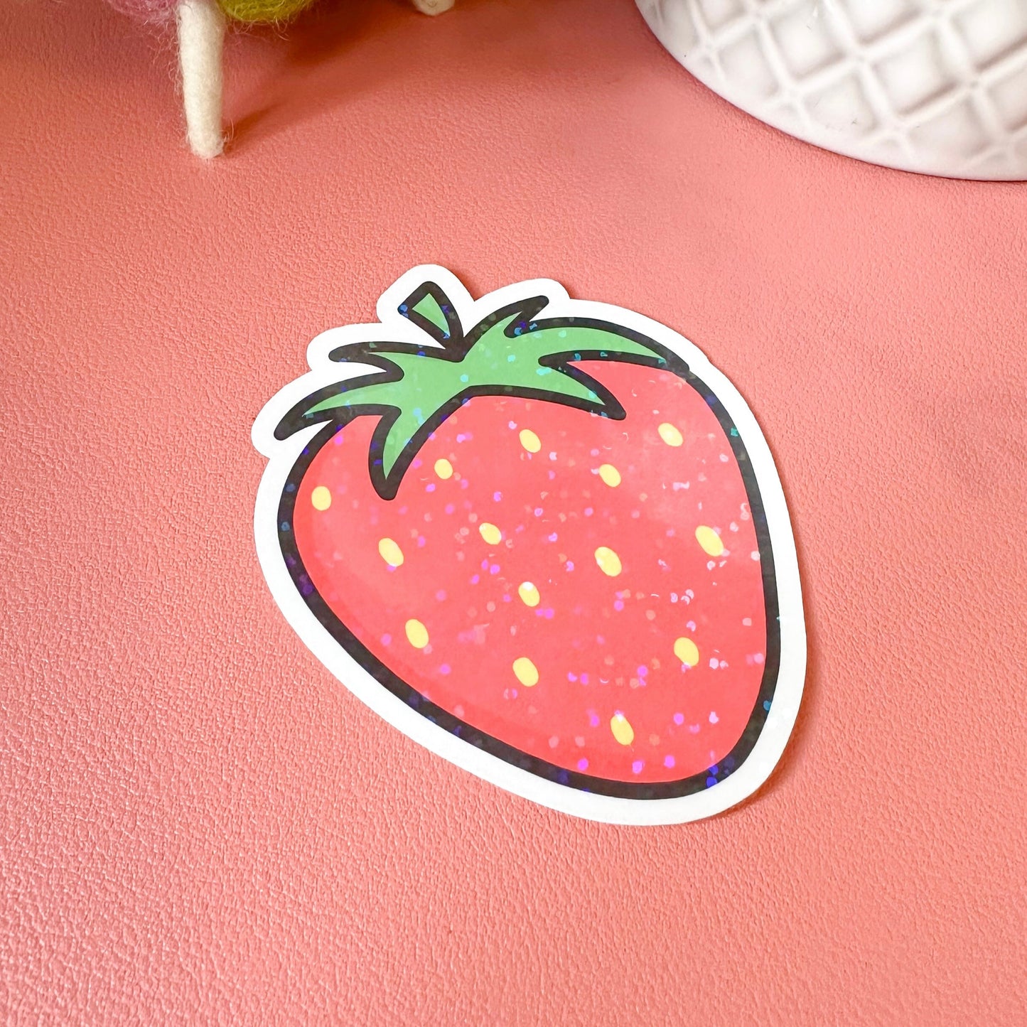 Strawberry-shaped sticker on a pink surface with decorative items in the background