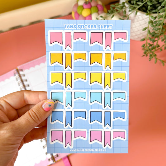 Sheet of colorful planner stickers held by a hand with a pink nail polish, on a pink surface with a plant in the background.