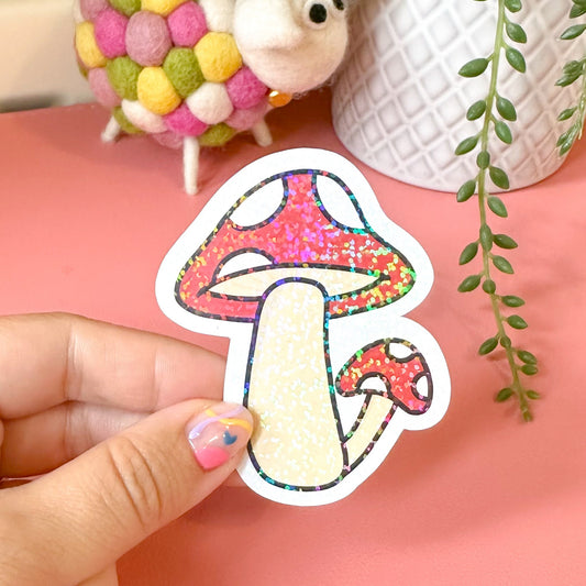 Hand holding a colorful mushroom sticker with a pink background, small plant, and decorative items.