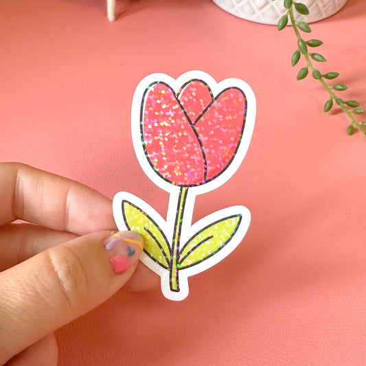Hand holding a pink tulip-shaped sticker against a pink background with decorative items.