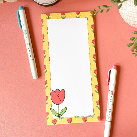 Notepad with a floral design on a pink surface with pens and plants.
