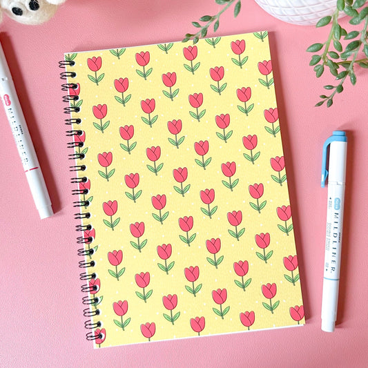 Notebook with pink tulip pattern on a pink surface with pens and plants.