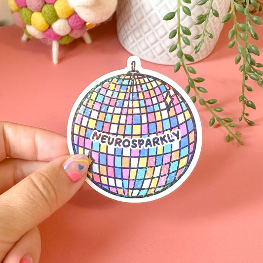 Colorful disco ball sticker with 'Neurosparkly' text held by a hand on a pink surface with decorative items.