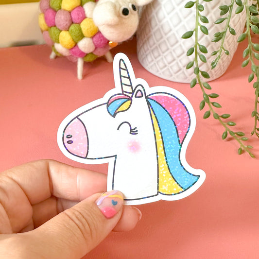 Hand holding a colorful unicorn sticker on a pink surface with decorative items in the background.
