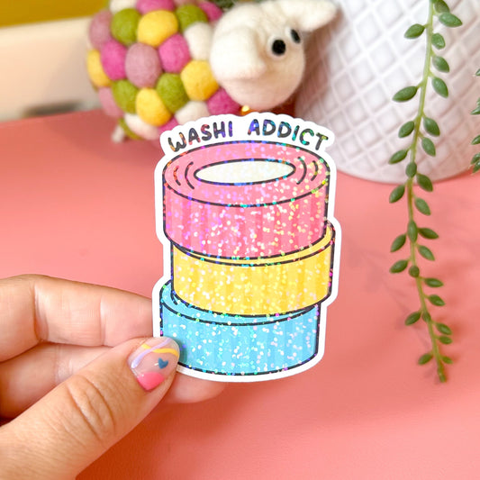 Sticker with colorful washi tape design held by a hand on a pink surface.