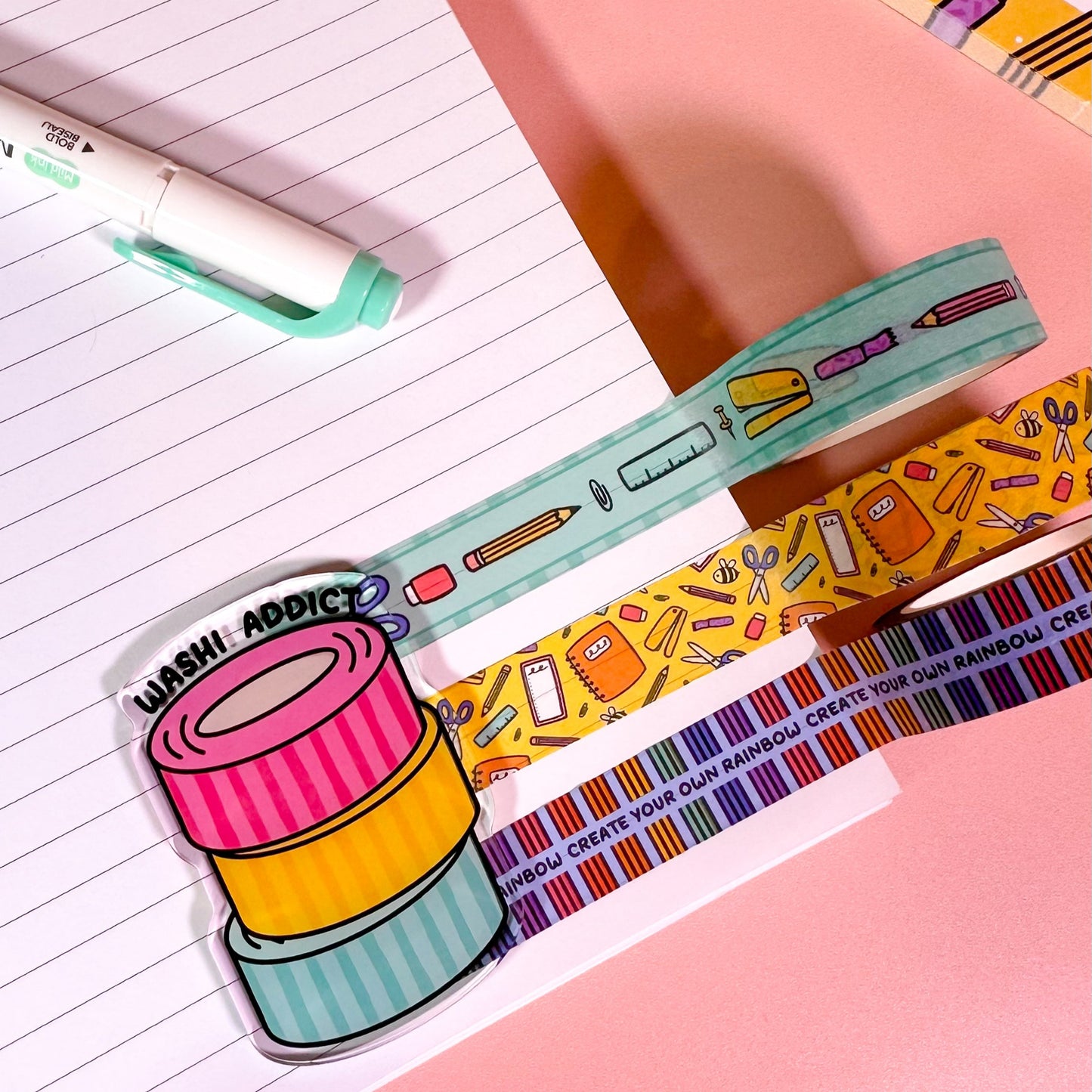 Colorful washi tape rolls on a pink surface with stationery items.