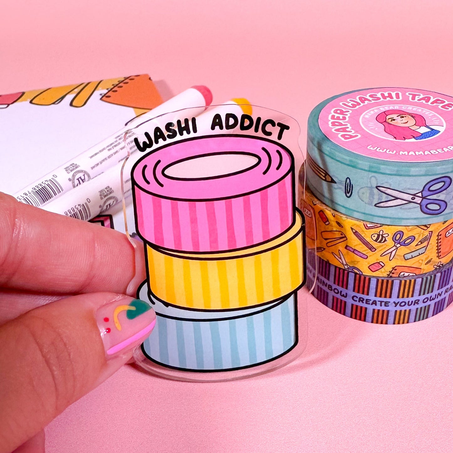 Stack of colorful washi tape with a hand holding one roll on a pink background