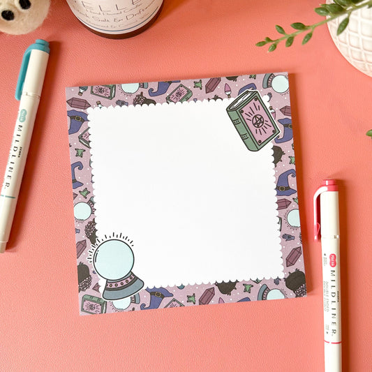 Notepad with witchy design on a pink surface with pens and a plant.