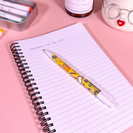 Notebook with a pen on a pink surface, surrounded by stationery items.